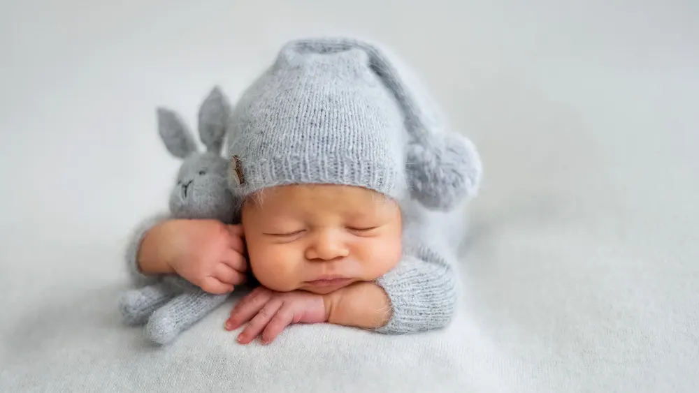Ein schlafendes Baby in grauer Strickkleidung und Mütze hält ein graues Hasenstofftier auf einer weißen Decke.