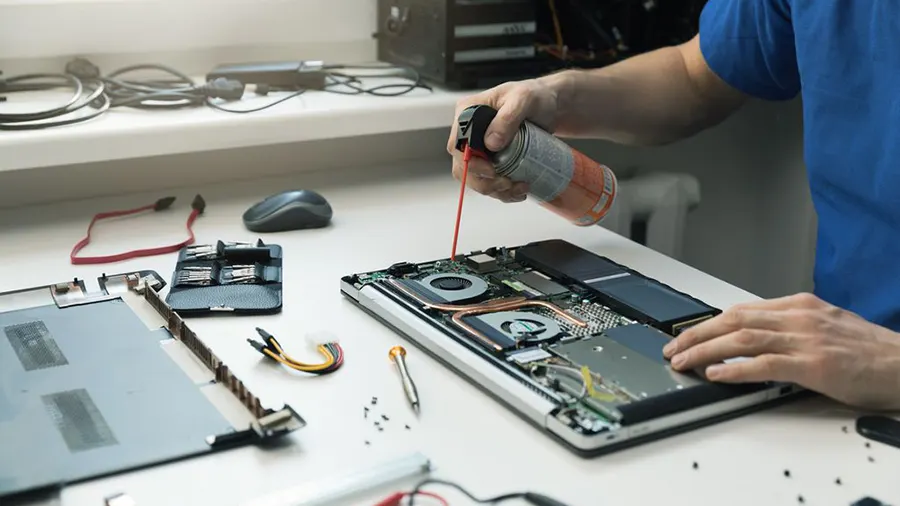 Eine Person reinigt einen geöffneten Laptop mit einer Dose Druckluft. Werkzeuge und Kabel liegen auf einem Tisch.