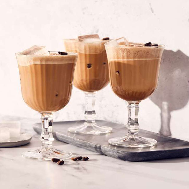 Drei Stielgläser mit braunem Getränk, Eiswürfeln und Kaffeebohnen auf einem grauen Tablett vor weißem Hintergrund.