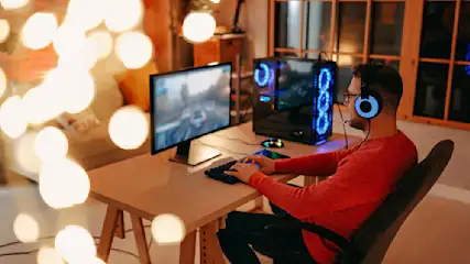 Ein Mann mit Kopfhörern sitzt vor einem Computer mit blau beleuchtetem Gehäuse und spielt ein Videospiel an einem hellbraunen Tisch.