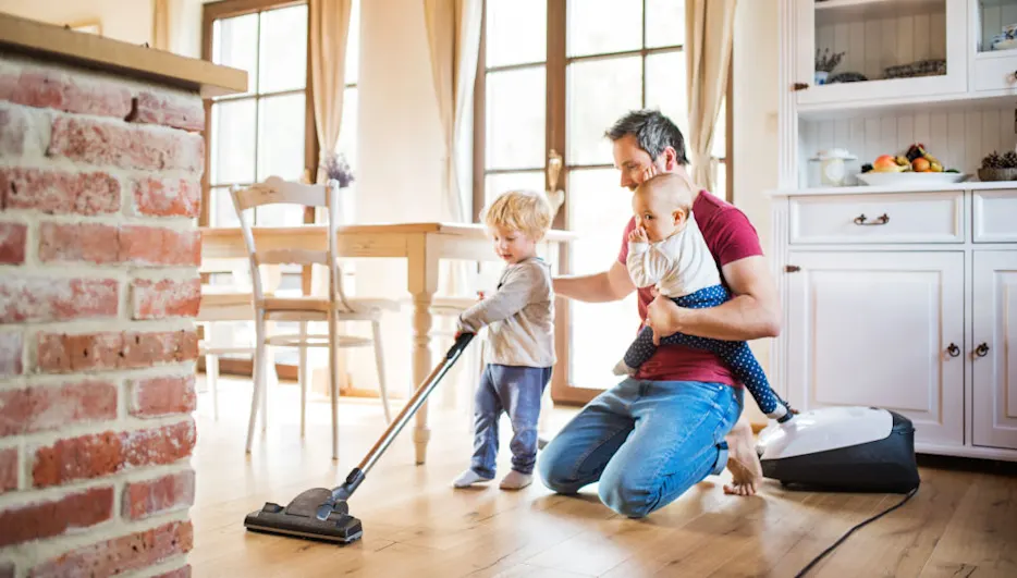 Ein Mann kniet mit einem Baby im Arm, während ein Kind mit einem Staubsauger auf einem Holzboden saugt.
