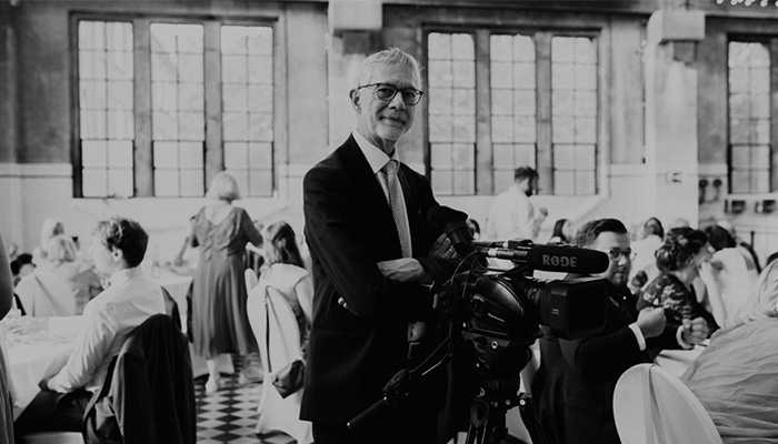 Ein Mann mit Brille und Anzug steht vor einer Videokamera in einem Raum mit Fenstern und anderen Personen.