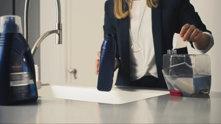 Frau gießt Flüssigkeit aus einer blauen Flasche in einen Behälter neben einem Waschbecken. Eine weitere blaue Flasche steht daneben.