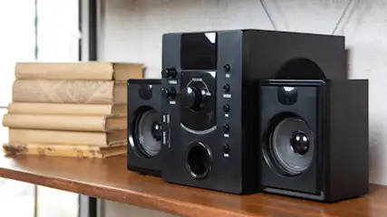 Auf einem Holzregal stehen Lautsprecher und ein Stapel Bücher. Die Lautsprecher sind schwarz und haben Regler für Lautstärke und Bass.