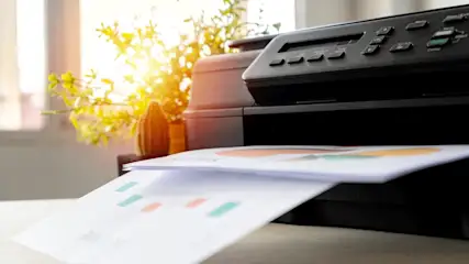 Ein schwarzer Drucker druckt ein Blatt Papier mit Diagrammen und farbigen Markierungen auf einem Tisch. Im Hintergrund steht eine Pflanze.