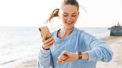 Eine Frau joggt am Strand mit Smartphone in einer Hand und schaut auf ihre Smartwatch.