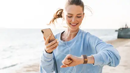 Frau mit blauem Pullover, Kopfhörern und brauner Uhr, die ein goldenes Handy am Strand mit Meeresblick hält.