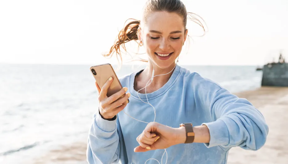Frau mit blauem Pullover, Kopfhörern und brauner Uhr, die ein goldenes Handy am Strand mit Meeresblick hält.