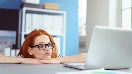 Eine Person mit Brille lehnt am Schreibtisch vor einem Laptop. Im Hintergrund befinden sich ein Regal und eine blaue Wand.
