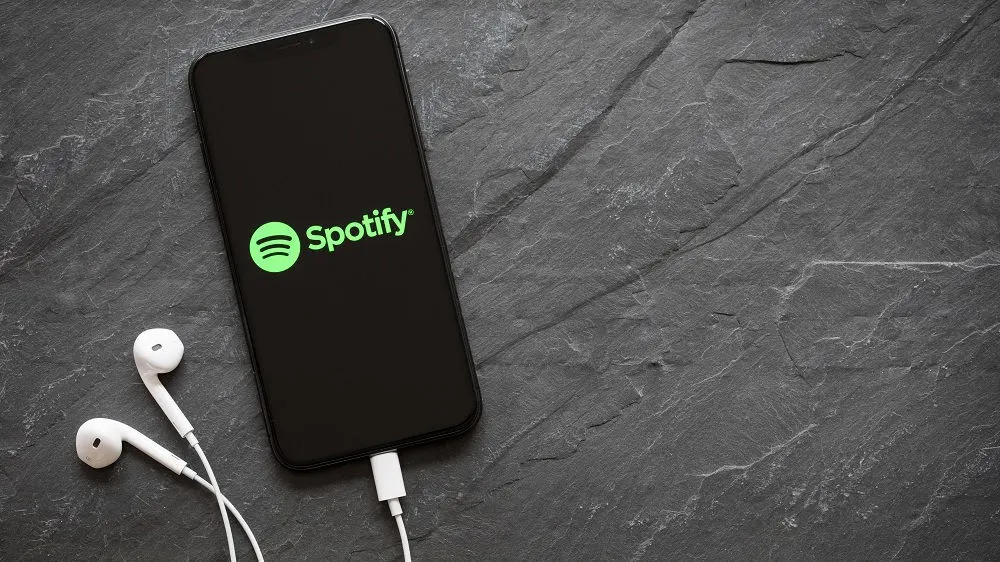 Ein Smartphone mit dem Spotify-Logo auf dem Display liegt auf einer schwarzen Oberfläche und ist mit weißen Kopfhörern verbunden.