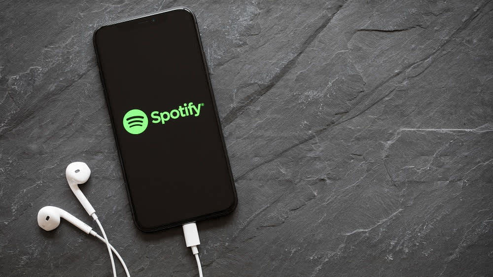 Ein Smartphone mit dem Spotify Logo auf dem Display liegt auf einer schwarzen Oberfläche und ist mit weißen Kopfhörern verbunden.