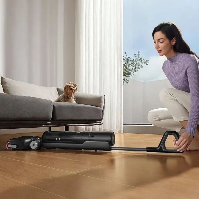 DREAME H12 Pro Flexlite Nass-/Trockensauger, Akkubetrieb Eine Frau schiebt den DREAME H12 FlexLite Nass-/Trockensauger flach über den Holzboden unter ein Sofa, auf dem Sofa sitzt ein kleiner Hund.