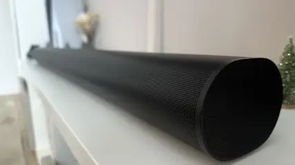 Ein schwarzer Lautsprecher liegt auf einer weißen Oberfläche. Im Hintergrund sind ein Fenster und eine kleine Dekoration zu sehen.