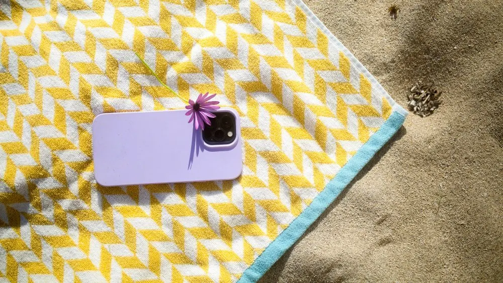 Handtuch mit gelbem Muster, lila Handy mit Blume und Sand im Hintergrund.
