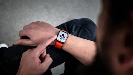 Eine Person mit einer orangefarbenen Smartwatch tippt auf das Display, mit Jeans im Hintergrund.