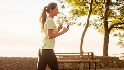 Eine Frau mit Pferdeschwanz hält ein Mobiltelefon. Sie trägt ein gelbes Oberteil und Leggings. Im Hintergrund befinden sich Bäume und eine Bank.