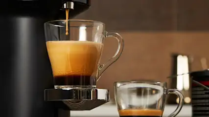 Espresso fließt in eine klare Tasse auf einer Maschine. Eine zweite Tasse steht daneben. Der Hintergrund ist unscharf.
