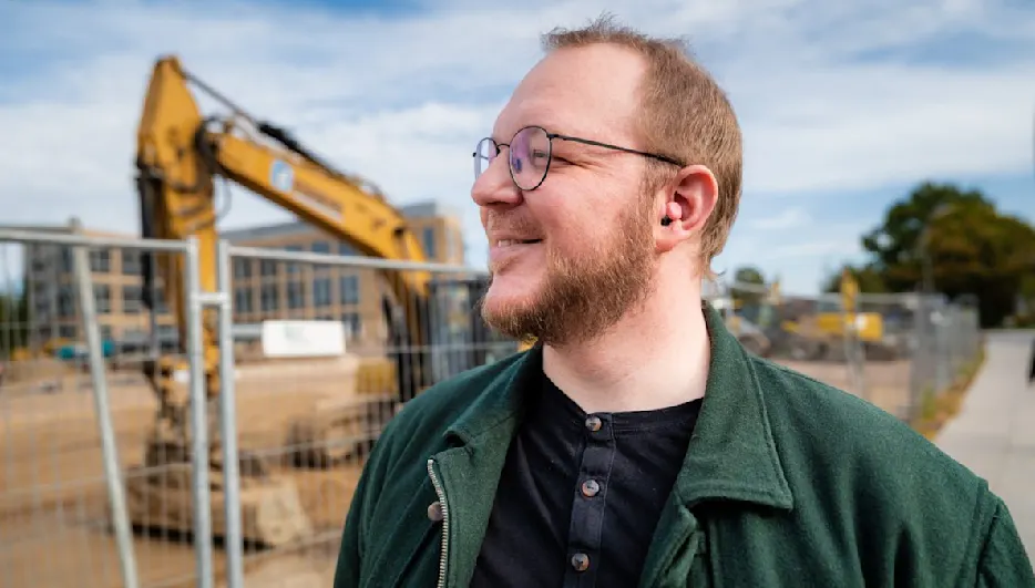 Ein Mann mit Brille und Bart lächelt im Vordergrund; im Hintergrund ein Bagger und ein Bauzaun unter blauem Himmel.