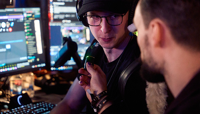 Mann mit Brille und Hut hält grünen Gegenstand vor Computermonitoren. Eine andere Person ist teilweise im Bild. Tastatur sichtbar.