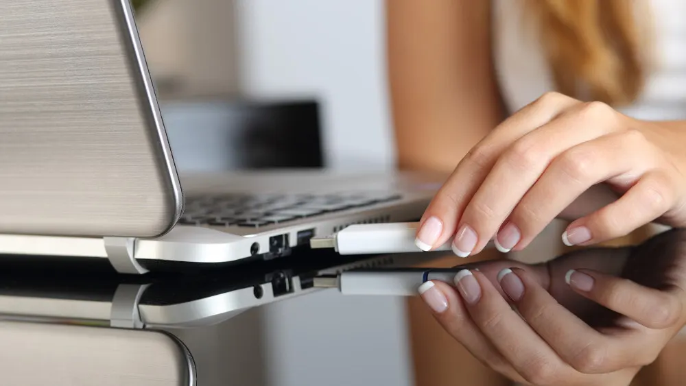 Eine Person steckt einen weißen USB-Stick in einen Laptop auf einer glänzenden Oberfläche.