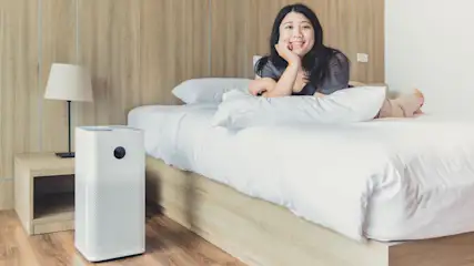 Eine Frau liegt auf einem Bett mit weißer Bettwäsche. Ein Luftreiniger steht auf einem Nachttisch neben einer Lampe.