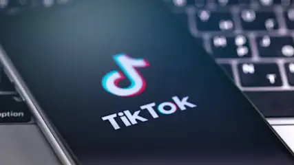 Ein Smartphone zeigt das TikTok-Logo vor einer unscharfen Computertastatur. Das Logo ist weiß mit blauem und rotem Rand.