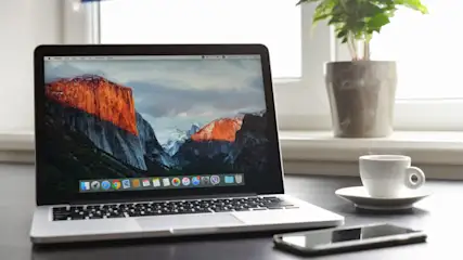 Ein Laptop zeigt ein Berglandschaft-Hintergrundbild. Daneben stehen eine Kaffeetasse, ein Smartphone und eine Topfpflanze vor einem Fenster.