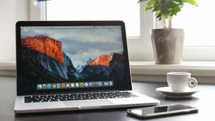 Ein geöffnetes MacBook, ein iPhone und eine weiße Tasse stehen auf einem Schreibtisch.