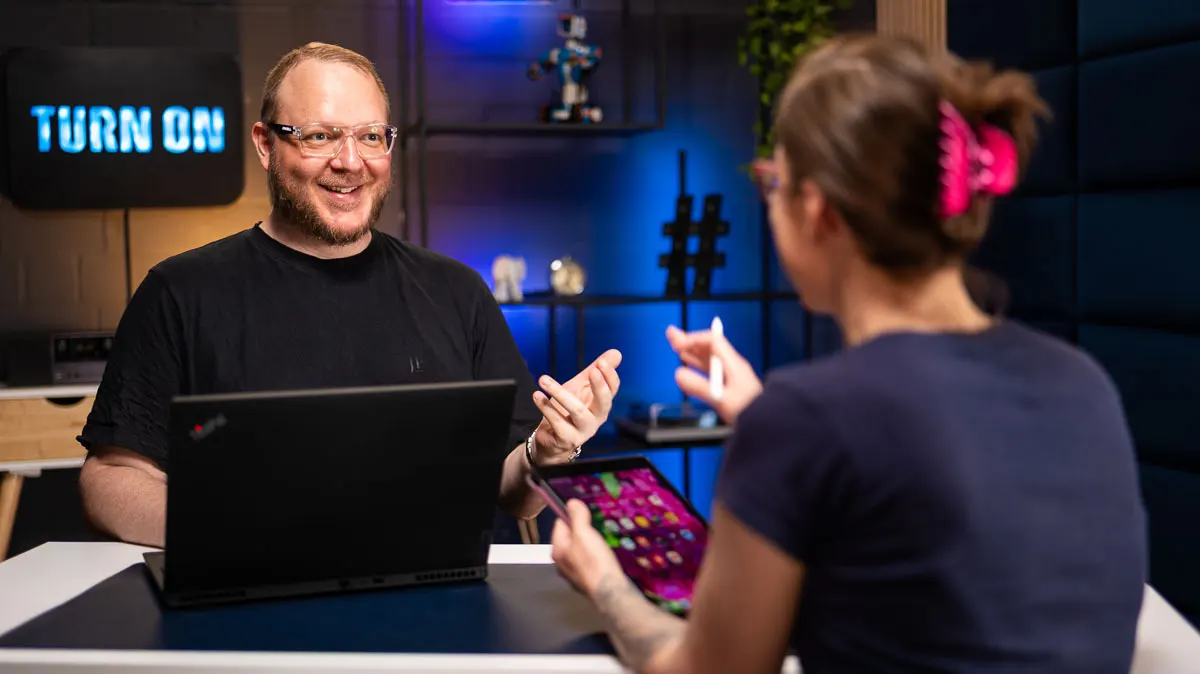 Andreas sitzt vor einem Laptop und schaut zu Daniela, die ein iPad in einer Hand und einen Apple Pencil in der anderen hält.