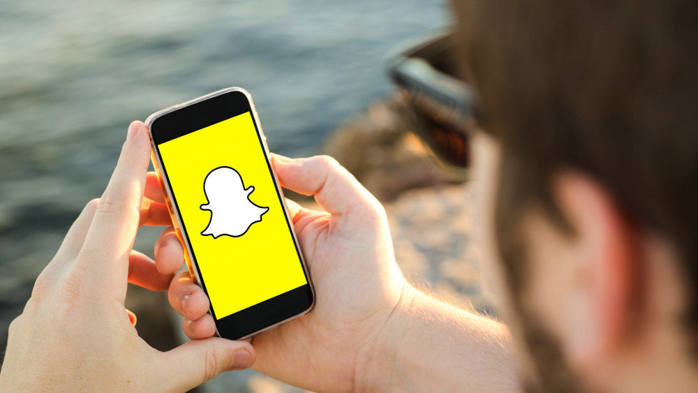 Eine Person hält ein Smartphone in der Hand, auf dessen Display das Snapchat-Logo abgebildet ist.
