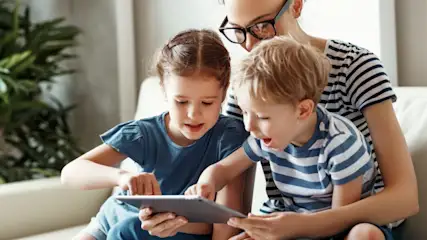Mutter mit zwei Kindern, die ein Tablet auf einem hellen Sofa benutzen. Die Mutter trägt eine gestreifte Bluse und eine Brille.