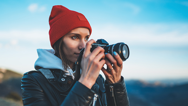 Eine Frau mit roter Mütze hält eine Kamera vor blauem Himmel. Sie trägt eine Jacke und einen Kapuzenpullover.