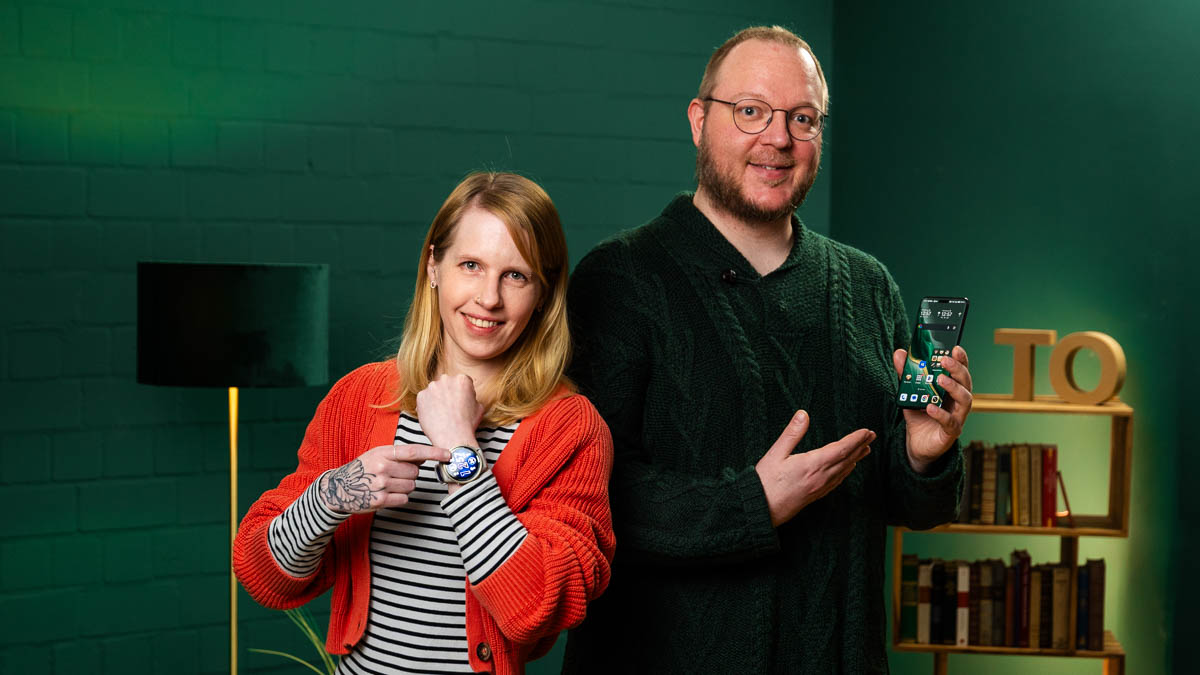 Franzi zeigt auf ihre Smartwatch und Andreas auf sein Smartphone.