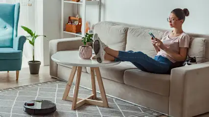 Eine Frau sitzt auf einem Sofa und benutzt ein Handy. Ein Staubsaugerroboter reinigt den Teppich im Wohnzimmer.