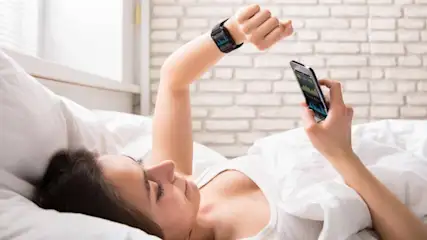 Eine Person liegt im Bett mit weißer Bettwäsche, betrachtet ein Handy und trägt eine schwarze Smartwatch. Ziegelwand im Hintergrund.