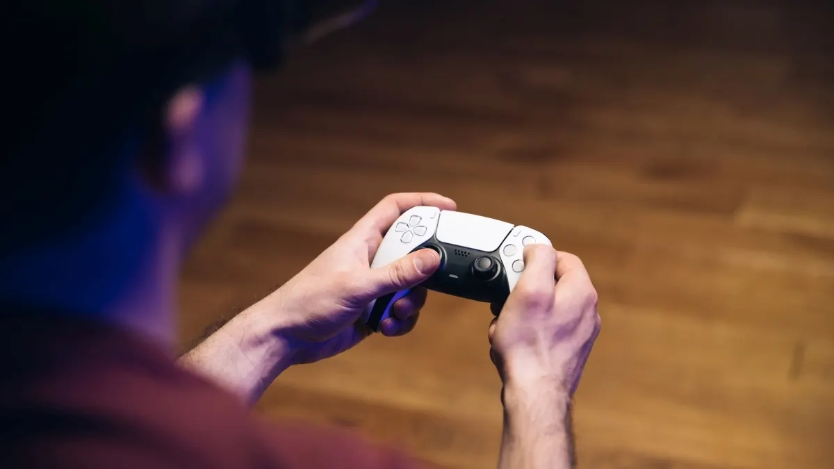 Eine Person hält einen weißen und schwarzen PlayStation 5 DualSense-Controller vor einem braunen Holzhintergrund.