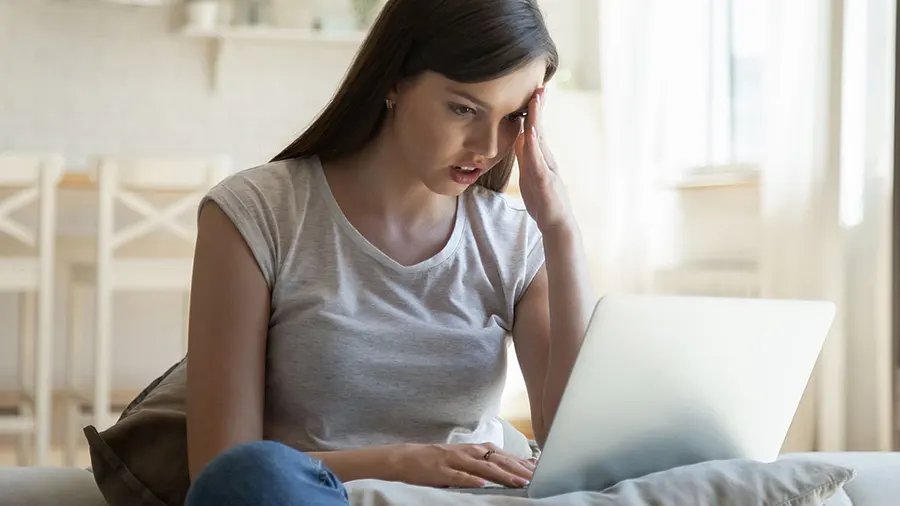 Eine Frau sitzt mit einem Laptop auf einem Sofa und hält eine Hand an ihre Stirn.