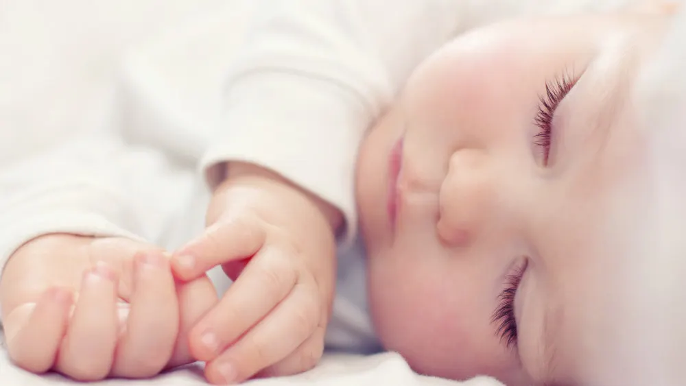 Ein schlafendes Baby liegt auf einem weißen Tuch und hält seine Hände vor dem Gesicht. Es trägt ein weißes Oberteil.