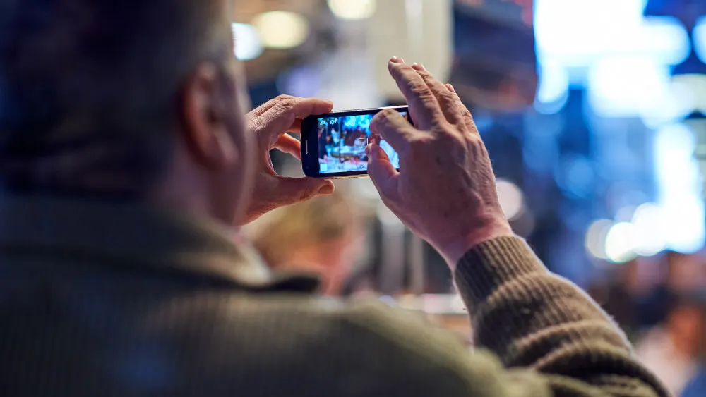 Eine Person mit beigem Pullover hält ein Smartphone horizontal, um etwas zu filmen. Im Hintergrund sind blaue Lichter.