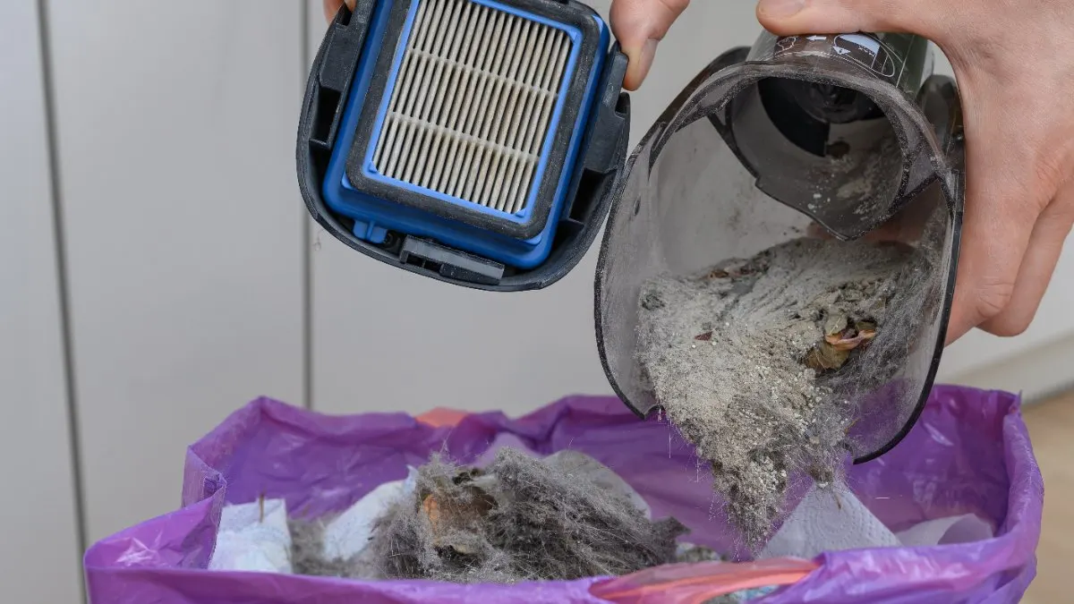 Eine Person leert den Staubbehälter eines Staubsaugers im Mülleimer aus.