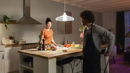 Eine Frau und ein Mann stehen an einer Kücheninsel mit Gemüse und Utensilien unter einer Lampe.