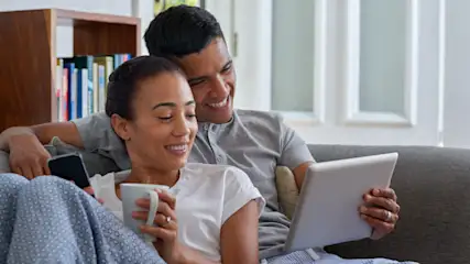 Ein Paar entspannt auf einem grauen Sofa; sie hält eine Tasse und ein Smartphone, er ein Tablet, Bücherregal im Hintergrund.
