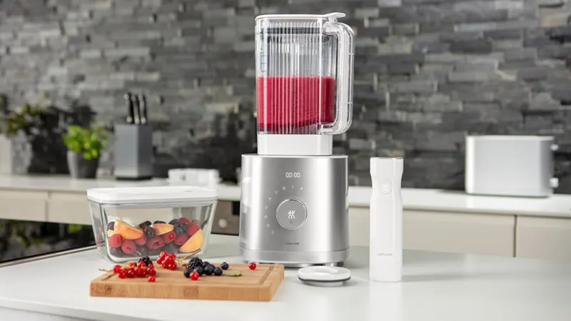 In der Küche steht ein Mixer mit rotem Inhalt. Daneben liegen Früchte auf einem Brett und in einer Schale.