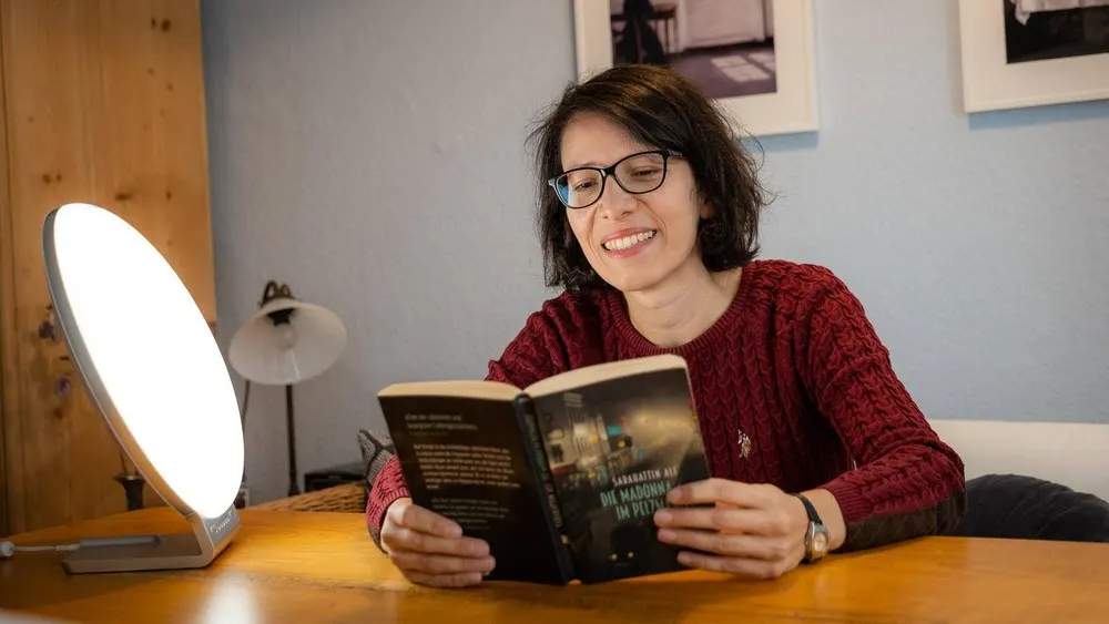 Frau mit Brille liest Buch am Tisch. Eine helle Lampe steht links. Im Hintergrund sind Bilder und eine Lampe sichtbar.
