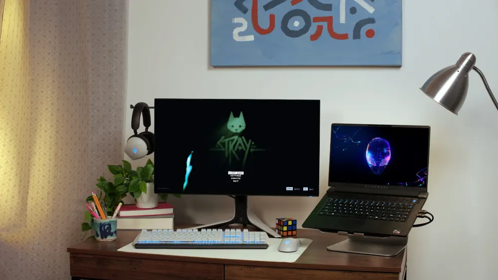 Ein Schreibtisch mit Monitor, Laptop, Tastatur, Maus und Rubik-Würfel unter einer Wandlampe. An der Wand hängt ein Bild.