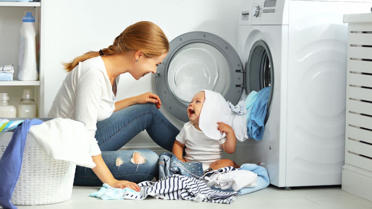 Eine Frau und ein Baby sitzen vor einer Waschmaschine und lachen, das Baby hat Wäsche auf dem Kopf.