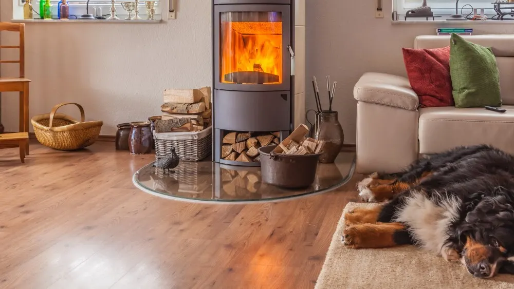 Ein Wohnzimmer mit Holzofen, Holzscheiten, Körben und einem schlafenden Hund auf einem Teppich vor einem hellen Sofa.