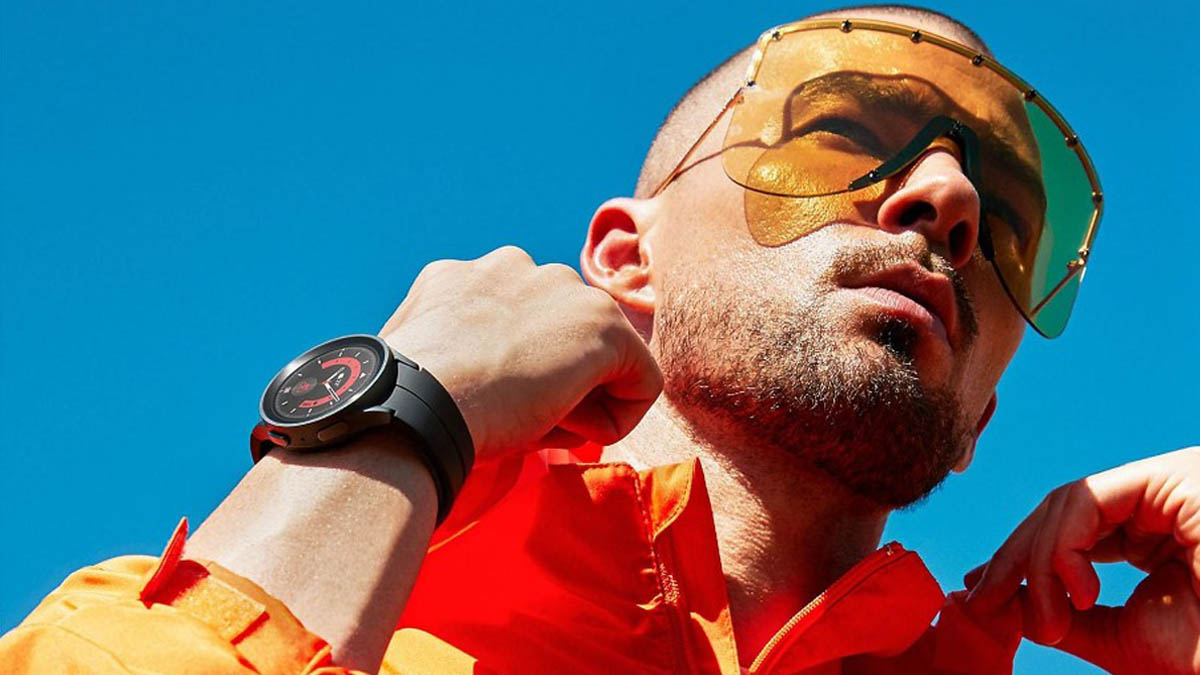 Mann mit gelber großer Sonnenbrille und orange-roter Jacke richtet sich den Kragen. An einer Hand sieht man die Samsung Galaxy Watch 5 Pro.