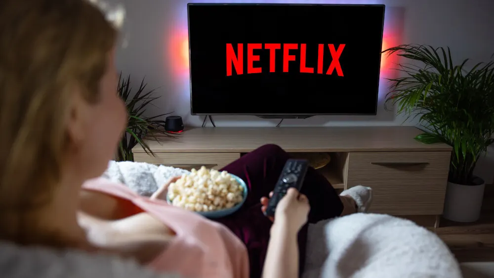 Eine Person liegt auf einem Sofa mit Popcorn und einer Fernbedienung vor einem Fernseher mit rotem Netflix-Logo.