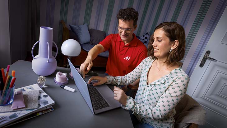 Zwei Personen arbeiten an einem Laptop an einem Schreibtisch mit Stiften, einer Vase und Zeitschriften in einem Raum.